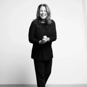 Kvinde i sort tøj smiler foran lys baggrund, hænder samlet. Sort/hvid billede.