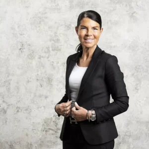 Kvinde i sort blazer og hvid top foran lys grå væg, med opsat hår og smilende udtryk.